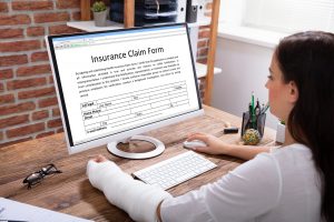 Businesswoman With A Plaster Cast Hand Filling Insurance Claim Form On Computer In The Office